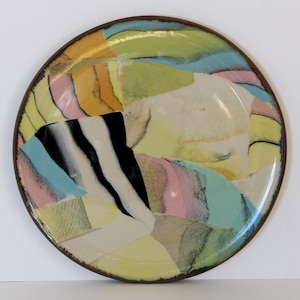 May include: A round ceramic plate with a brown rim and a colorful abstract design. The design features patches of yellow, pink, blue, orange, and black, creating a patchwork effect. The plate has a glossy finish.