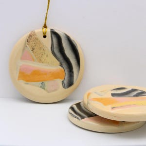 May include: Three round ceramic ornaments with a beige background and abstract designs in black, pink, and yellow.
