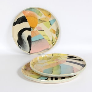 May include: Three round ceramic plates featuring an abstract collage design. The surface displays irregular shapes in shades of black, orange, yellow, light green, blue, and pink against an off-white background with a glossy finish.