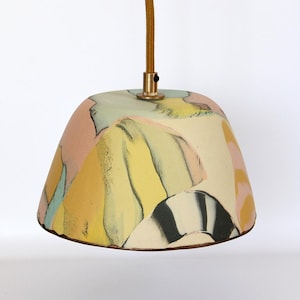 May include: A ceramic pendant light with a colorful, abstract design. The lampshade features a mix of pastel colors, including pink, yellow, and blue, with black and white striped accents. It hangs from a gold-colored cord.