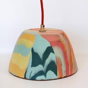 May include: A dome-shaped pendant lamp shade featuring a marbled pattern in shades of yellow, teal, dark green, and coral. The shade is suspended by a bright orange fabric cord attached to a small brass-coloured metal fixture at the top centre.