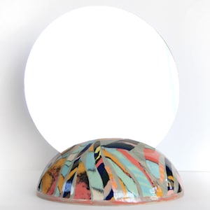 Puede incluir: Un cuenco redondo de cerámica con un diseño abstracto colorido. El cuenco está pintado en tonos de azul, verde, amarillo, naranja y rosa. El cuenco está sobre una superficie blanca.