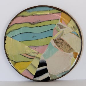 May include: A round ceramic plate with a brown rim. The plate features a colorful abstract design with pastel pink, blue, yellow, and green sections. The design includes black and white striped elements.
