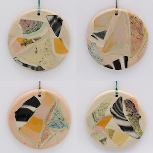 May include: Four round ceramic ornaments with a white background and a colorful abstract design. The ornaments are decorated with pink, yellow, black, and blue geometric shapes.