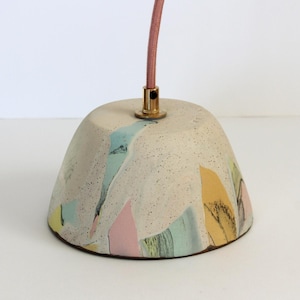 May include: A speckled, off-white ceramic pendant light fixture with colorful abstract shapes in pastel hues. The light has a pink fabric-covered cord and a gold-colored metal fitting. The fixture is a unique, artistic home decor item.