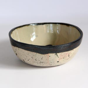 Puede incluir: Un cuenco de cer&aacute;mica con interior crema y exterior moteado blanquecino. El borde es una banda oscura y texturizada. El cuenco tiene una forma ligeramente irregular y es adecuado para servir alimentos.