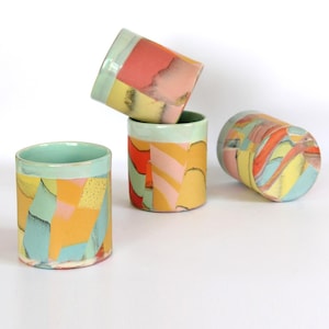 May include: Four cylindrical ceramic cups featuring a multi-colored abstract pattern of yellow, coral, light blue, and pink shapes. The cups have a smooth finish with some speckled textures visible on the surface, arranged in a cluster.
