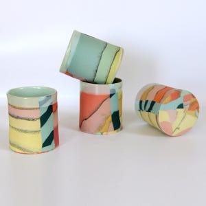 Puede incluir: Cuatro vasos de cerámica con diseños abstractos. Los vasos presentan una mezcla de colores, incluyendo amarillo, rosa, naranja, verde azulado y negro. Los vasos son cilíndricos con un borde ligeramente ensanchado. Un vaso está tumbado de lado.
