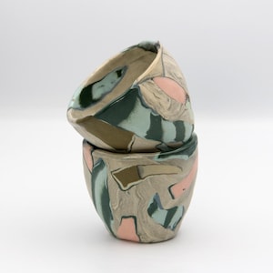 May include: Two stacked ceramic cups with abstract designs. The cups feature a beige base color with green, pink, and blue organic shapes. The cups have a unique, asymmetrical shape, with a slightly tilted rim.
