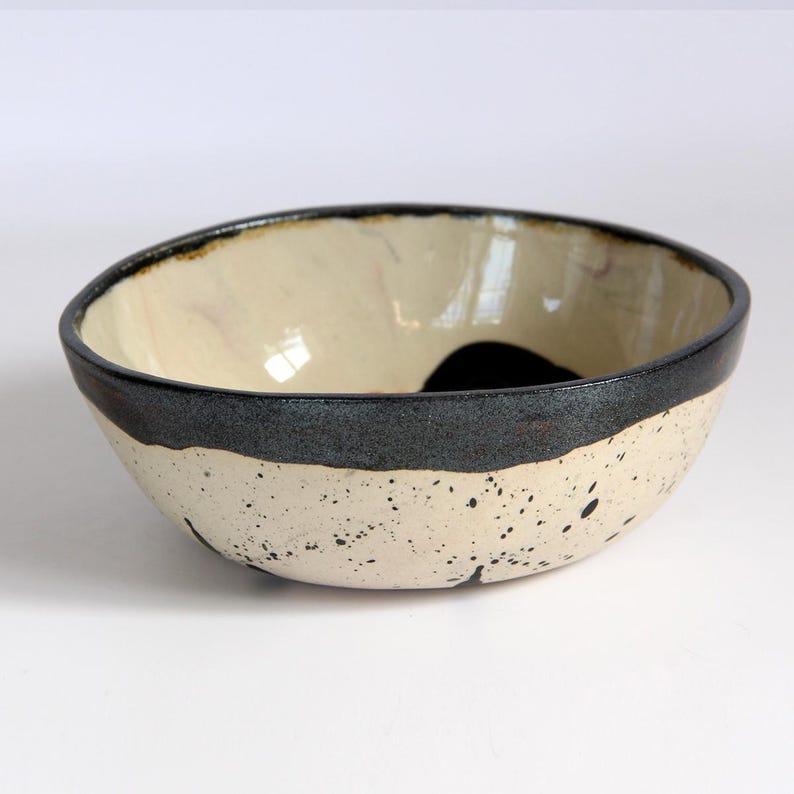 Puede incluir: Un cuenco de cer&aacute;mica con interior crema y exterior moteado blanquecino. Una gruesa banda gris oscuro rodea el cuenco, con un acento negro en el interior. El cuenco tiene una forma ligeramente irregular.
