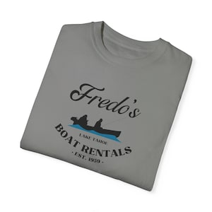May include: Gray t-shirt with a black and white graphic of a boat with two people fishing. The text "Fredo's Boat Rentals. Est. 1959." is printed below the graphic.