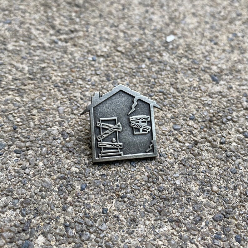 May include: A silver metal house pin with a boarded up door and a window with a boarded up pane. The house has a crack in the side.