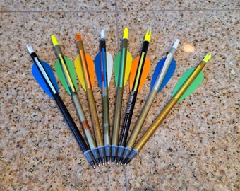 May include: A collection of ten colorful arrows with fletching in various colors, including blue, green, yellow, orange, and white. The arrows are arranged in a fan shape on a brown speckled surface.