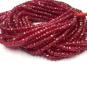 Pode incluir: Um fio de contas de rubi vermelho escuro, facetadas e polidas, dispostas em espiral sobre um fundo branco.