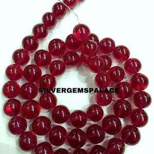 Op de afbeelding: Een streng ronde, rode edelsteen kralen. De kralen zijn doorschijnend en hebben een lichte glans. De kralen zijn geregen op een zilveren draad.