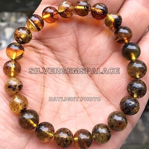 May include: A bracelet made of round, amber-coloured beads with dark brown inclusions. The beads are strung on a clear elastic cord. The bracelet is on a person's wrist. The text "SILVERGEMSPALACE DAYLIGHT PHOTO" is visible in the image.