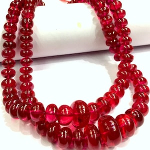 Op de afbeelding: Een streng diepe rode robijn kralen, elke kraal is rond en licht langwerpig, waardoor een textuur ontstaat. De kralen zijn aan elkaar geregen om een lange ketting te vormen.