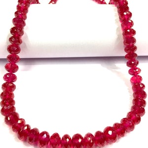 May include: A strand of deep red ruby beads, each bead is faceted and has a slightly elongated shape. The beads are strung on a clear thread and are arranged in a simple, elegant design.