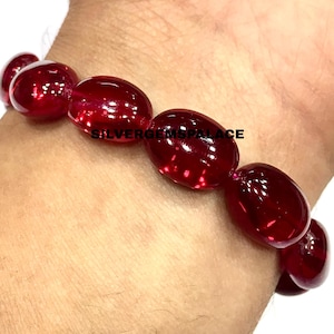 Op de afbeelding: Een armband bestaande uit gladde, ovaalvormige, doorschijnende dieprode kralen. De kralen hebben een gepolijst, reflecterend oppervlak. De tekst SILVERGEMSPALACE is in het zwart over het midden van de kralen gedrukt. De armband wordt om de pols gedragen.