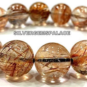 May include: A bracelet made of 12 round, clear, faceted beads with red and gold inclusions. The beads are strung on a silver chain. The bracelet is labeled "SILVERGEMSPALACE".