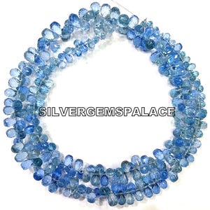 May include: A strand of light blue gemstone beads, with a gradient of color from dark to light blue. The beads are teardrop shaped and strung on a clear thread. The beads are arranged in a circle, with the ends of the strand overlapping. The text "SILVERGEMSPALACE" is visible in the center of the image.