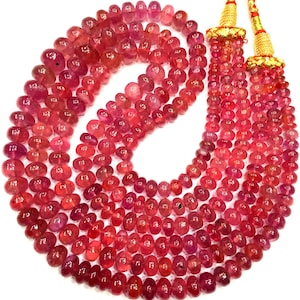 Può includere: Una collana di perle di rubino rosa con un fiocco dorato. Le perle sono rotonde e hanno una finitura liscia e lucida.