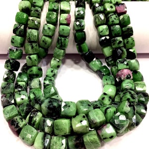 May include: A strand of green gemstone beads with black and white inclusions. The beads are faceted and have a square shape.
