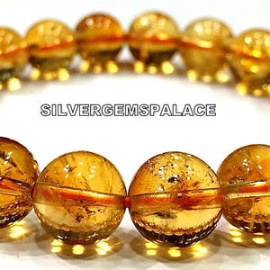 Peut inclure: Un bracelet composé de perles de citrine dorées avec une bande rouge traversant chaque perle. Le bracelet est disposé sur une surface blanche. Le texte "SILVERGEMSPALACE" est visible au centre de l'image.