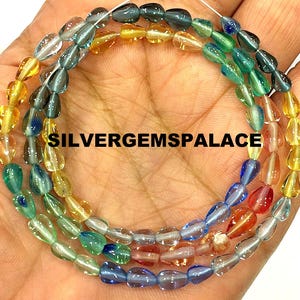 Op de afbeelding: Slingers van kleurrijke, druppelvormige kralen in tinten geel, groen, blauw en rood. De kralen zijn aan elkaar geregen en de tekst "SILVERGEMSPALACE" is zichtbaar. De kralen worden in een hand gehouden.