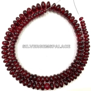 May include: A strand of deep red gemstone beads, likely ruby, with a round, slightly elongated shape. The beads are strung on a thin wire and are arranged in a circular pattern. The image includes the text "SILVERGEMSPALACE".
