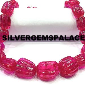Puede incluir: Un collar de cuentas de piedras preciosas de color rosa intenso y translúcidas. Cada cuenta tiene forma oblonga redondeada con detalles tallados y acanalados. El texto "SILVERGEMSPALACE" está impreso en la imagen. Las cuentas están ensartadas, lo que sugiere un collar o una pulsera.
