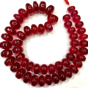 Puede incluir: Una hebra de cuentas de vidrio rojo oscuro, cada cuenta es redonda y ligeramente alargada, creando un aspecto único y llamativo. Las cuentas están enhebradas en un hilo transparente, creando un bucle continuo.