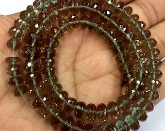 May include: Strand of faceted, teardrop-shaped beads in shades of brown and amber. The beads are strung on a light green thread, creating a layered effect. The beads are held in a hand.