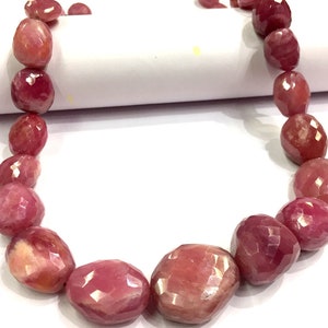 Könnte beinhalten: Eine Kette aus rosa Rubin-Edelsteinen, facettiert und oval geformt. Die Perlen sind auf einer weißen Schnur aufgefädelt und in einem Kreis angeordnet.