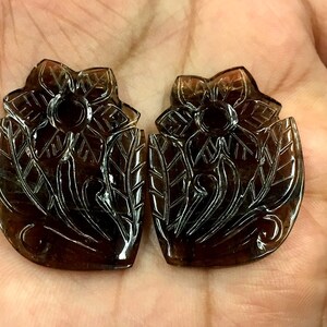 May include: Two brown gemstone flower carvings. The carvings are detailed and intricate, with a floral design. The gemstones are translucent and have a dark brown color.