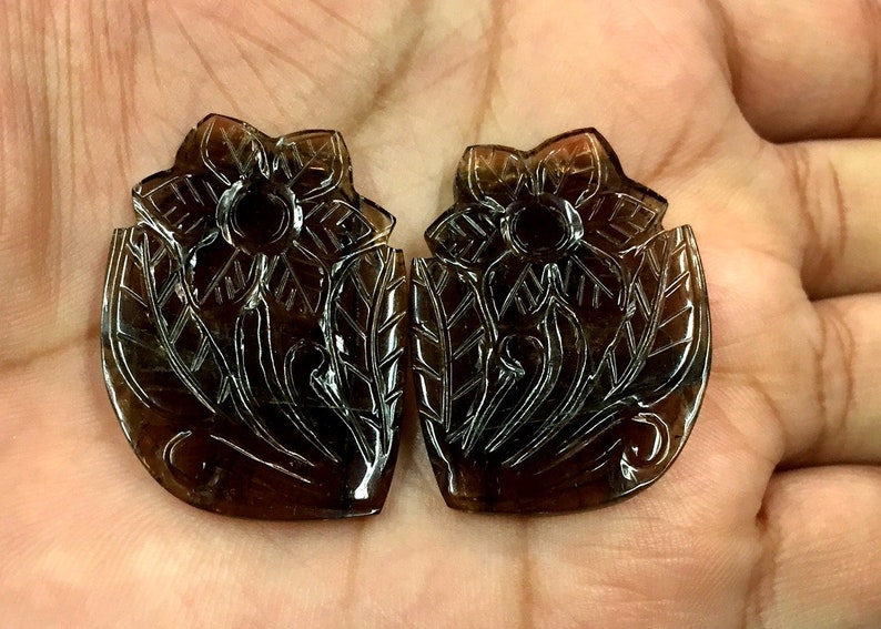 May include: Two brown gemstone flower carvings. The carvings are detailed and intricate, with a floral design. The gemstones are translucent and have a dark brown color.