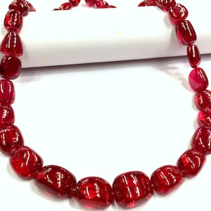 May include: A necklace made of bright red gemstone beads. The beads are faceted and have a rounded, slightly irregular shape. The necklace is strung on a clear thread.
