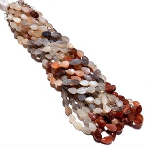 May include: A strand of gemstone beads in various shades of peach, gray, and red. The beads are pear-shaped and have a smooth, polished finish.