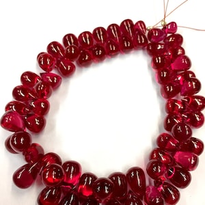 May include: A strand of deep red ruby gemstone beads in a teardrop shape. The beads are strung on a thin wire and are arranged in a circular pattern.