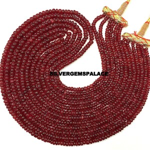 Può includere: Un filo di perle di rubino rosso scuro, di circa 4 mm di diametro, infilate su un cordoncino. Le perle sono disposte in un motivo circolare, creando una collana o un braccialetto.