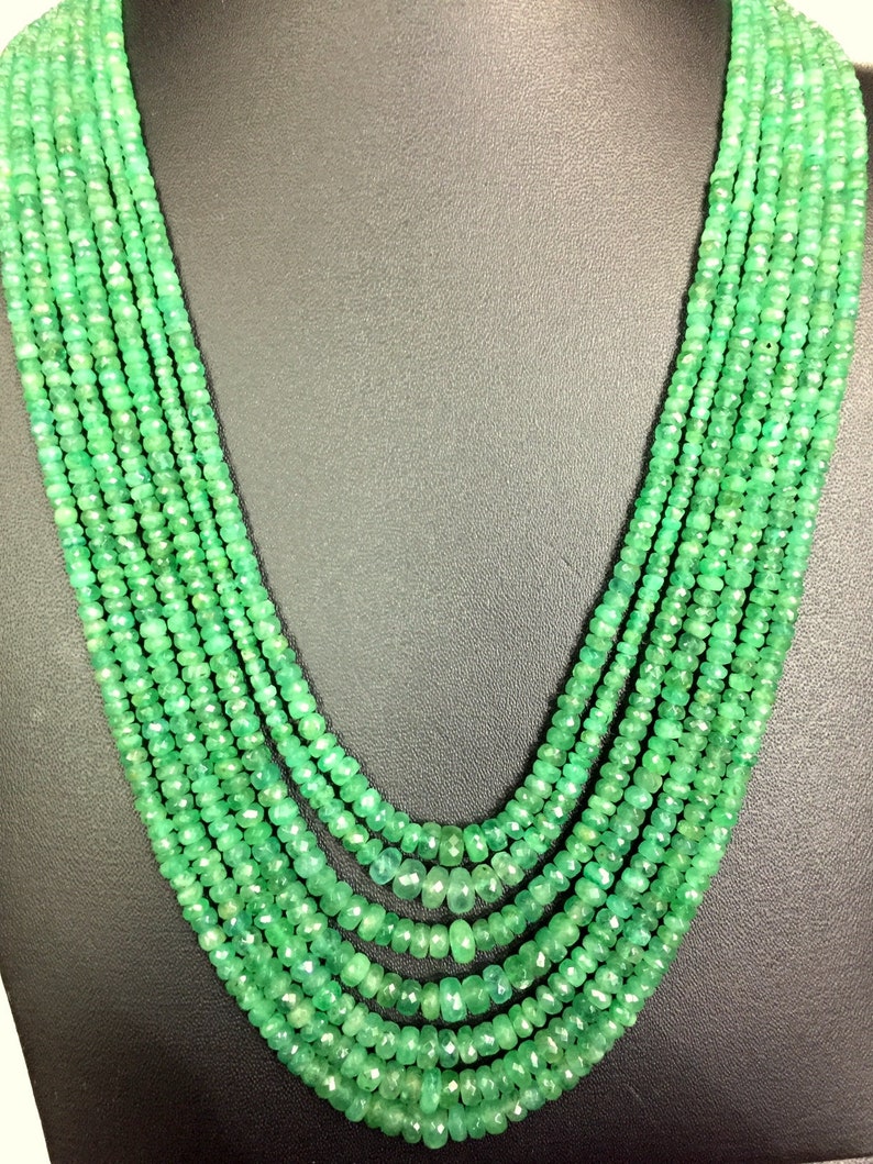 K&ouml;nnte beinhalten: Eine mehrstr&auml;ngige Halskette aus kleinen, facettierten, smaragdgr&uuml;nen Perlen. Die Halskette wird vor einem dunklen Hintergrund pr&auml;sentiert, der die lebendige Farbe und das aufwendige Design des Schmucks hervorhebt.