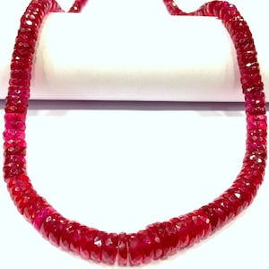 May include: A strand of deep red ruby beads, each bead is faceted and has a slightly irregular shape. The beads are strung on a clear thread and are arranged in a continuous loop.