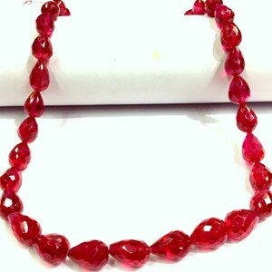 May include: A strand of deep red faceted gemstone beads in a teardrop shape. The beads are strung on a clear thread and are arranged in a circular pattern.