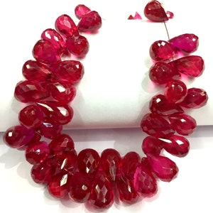 Puede incluir: Una hebra de cuentas de piedras preciosas facetadas de color rojo intenso en forma de lágrima. Las cuentas están enhebradas en un alambre fino y están dispuestas en un patrón suelto y fluido.