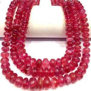 Puede incluir: Una hebra de cuentas de rubí rojo intenso, cada cuenta es lisa y redonda, con una forma ligeramente ovalada. Las cuentas están enhebradas en un hilo transparente, creando un collar hermoso y elegante.