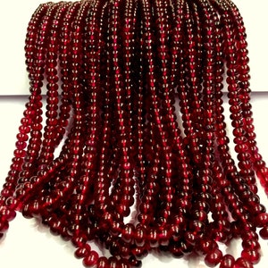 Op de afbeelding: Een streng dieprode edelsteen kralen. De kralen zijn rond en glad en aan elkaar geregen op een doorzichtige draad. De kralen zijn allemaal even groot en even gevormd en zijn in een nette, gelijkmatige rij gerangschikt.