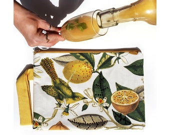 Puede incluir: Un bolso de mano blanco con un estampado botánico que presenta un pájaro verde, limones amarillos y maracuyá. Se está llenando un vaso con un líquido amarillo. El bolso de mano tiene una cremallera dorada y un asa amarilla.