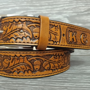 May include: A tan leather belt featuring an embossed pattern of oak leaves and acorns throughout the strap. The belt includes a silver-toned metal buckle and a leather keeper loop. The pointed end of the belt is stamped with the letters KC.