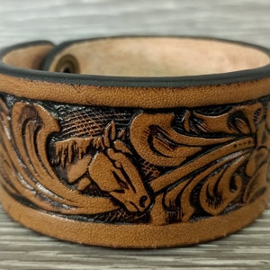 May include: A brown leather cuff bracelet with a horse head and floral design. The cuff is adjustable and features a detailed embossed pattern with dark brown accents. The edges are finished with black trim.