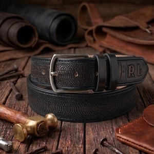 May include: A black leather belt with a silver buckle, featuring a textured pattern. The belt is stacked on top of another black leather belt. The initials "RL" are embossed on the belt. The image also includes leather rolls, tools, and a leather wallet.
